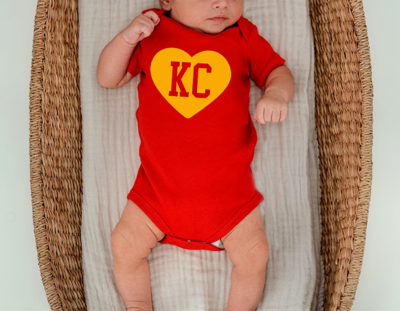May include: A baby wearing a red onesie with a yellow heart that says "KC" on it. The baby is lying in a wicker basket.