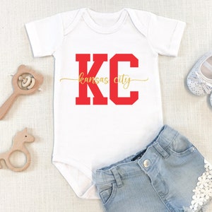 May include: White baby onesie with red "KC" lettering and gold "kansas city" text. The onesie is paired with denim shorts with white lace trim.