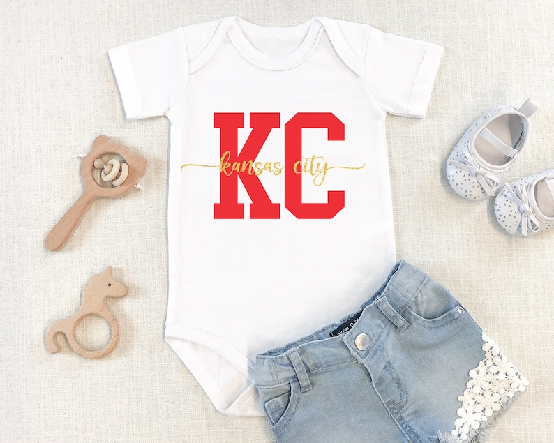 May include: White baby onesie with red "KC" lettering and gold "kansas city" text. The onesie is paired with denim shorts with white lace trim.