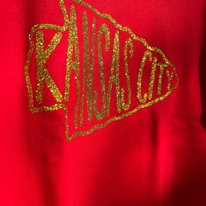 May include: Red sweatshirt with a gold glitter design of an arrow with the words "KANSAS CITY" inside.
