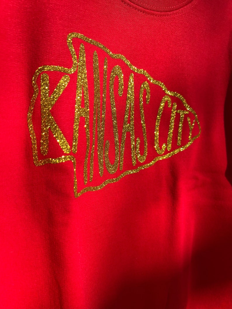 May include: Red sweatshirt with a gold glitter design of an arrow with the words "KANSAS CITY" inside.
