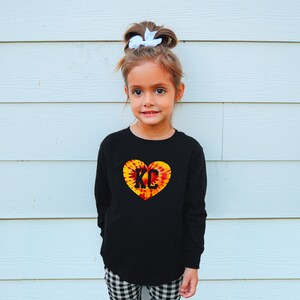 May include: A young girl wearing a black long-sleeved shirt with a tie-dye heart design featuring the letters "KC" in black. She has a white bow in her hair and is standing in front of a white wall.