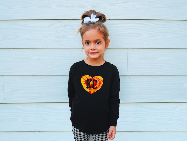 May include: A young girl wearing a black long-sleeved shirt with a tie-dye heart design featuring the letters "KC" in black. She has a white bow in her hair and is standing in front of a white wall.