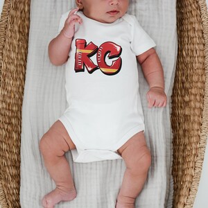 May include: A white baby onesie with a red and yellow graphic of the letters "KC" on the front. The onesie is worn by a baby lying in a wicker basket.