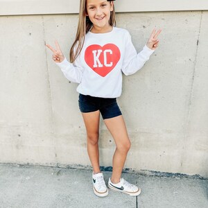 Peut inclure: Une jeune fille portant un t-shirt blanc &agrave; manches longues avec un motif de c&oelig;ur rouge sur lequel est inscrit "KC" en lettres blanches. Elle porte un short noir et des baskets blanches avec des accents noirs. Elle est debout devant un mur blanc.