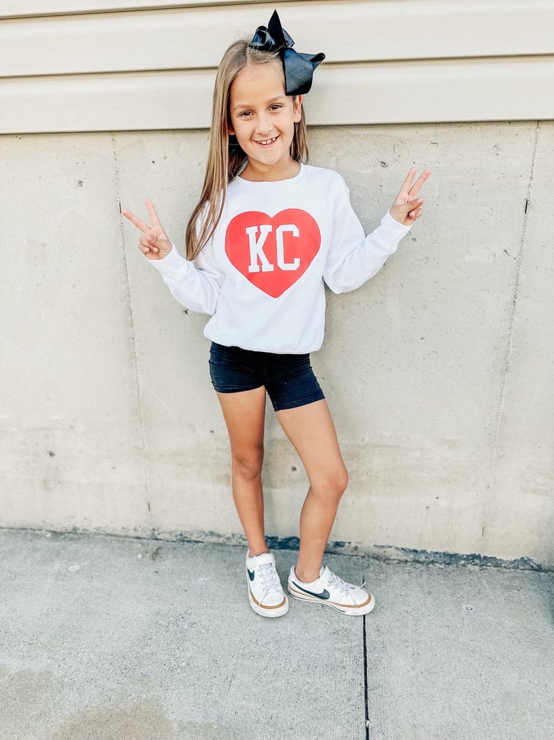 Peut inclure: Une jeune fille portant un t-shirt blanc &agrave; manches longues avec un motif de c&oelig;ur rouge sur lequel est inscrit "KC" en lettres blanches. Elle porte un short noir et des baskets blanches avec des accents noirs. Elle est debout devant un mur blanc.