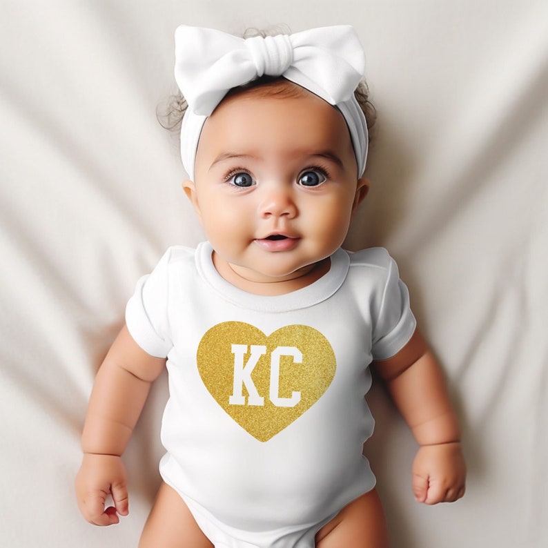 May include: A white baby onesie with a gold glitter heart that says "KC" on it. The baby is wearing a white headband with a bow.