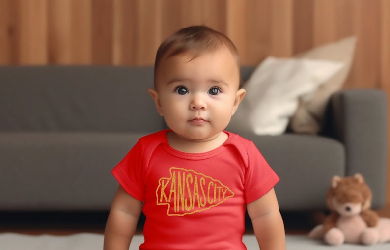 May include: A baby wearing a red onesie with a gold "Kansas City" arrow graphic. The baby is sitting on a white rug in front of a couch.