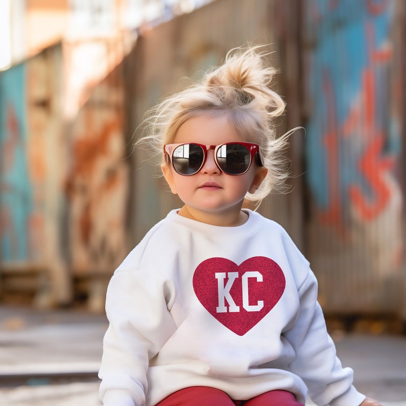 Peut inclure: Une jeune fille portant des lunettes de soleil rouges et un sweat-shirt blanc avec un c&oelig;ur rouge paillet&eacute; qui dit "KC" dessus.
