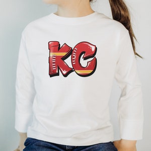 May include: White long-sleeve shirt with a red and yellow graphic of the letters "KC" designed to look like a football.