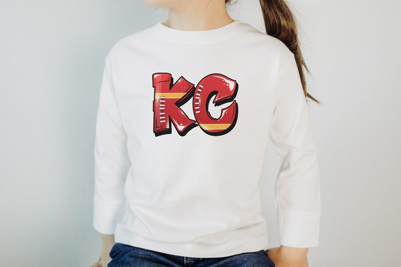 May include: White long-sleeve shirt with a red and yellow graphic of the letters "KC" designed to look like a football.