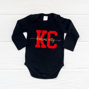 May include: Black long-sleeve baby onesie with red "KC" lettering and gold "kansas city" text.