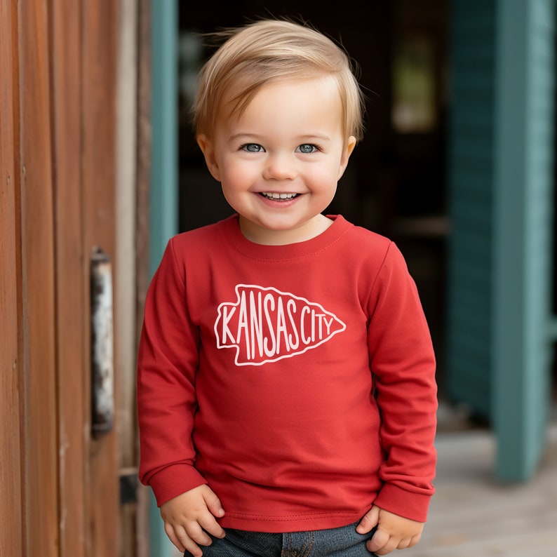 May include: A young child wearing a red long-sleeved shirt with a white graphic of an arrowhead with the text "KANSAS CITY" inside. The child is smiling and looking at the camera.