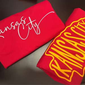 May include: Two red t-shirts with the text "Kansas City" in white and yellow lettering. One shirt has the text in a script font and the other has the text in a bold, blocky font.