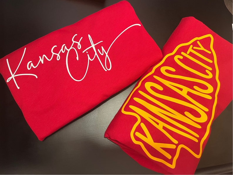 May include: Two red t-shirts with the text "Kansas City" in white and yellow lettering. One shirt has the text in a script font and the other has the text in a bold, blocky font.