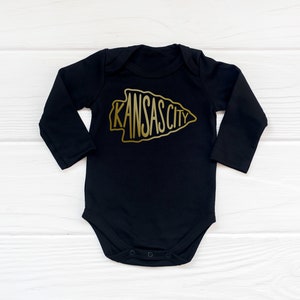May include: Black long-sleeve baby onesie with a gold arrowhead design featuring the text "KANSAS CITY".