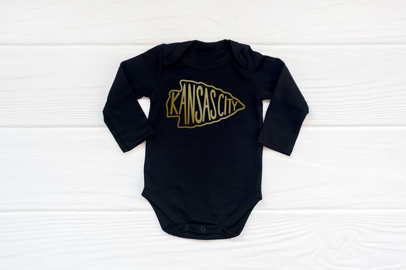 May include: Black long-sleeve baby onesie with a gold arrowhead design featuring the text "KANSAS CITY".