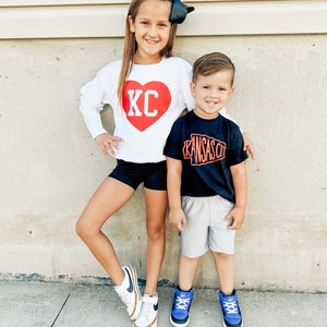 Peut inclure: Deux enfants portant des chemises assorties sur le th&egrave;me de Kansas City. La fille porte un sweat-shirt blanc avec un c&oelig;ur rouge sur lequel est inscrit "KC". Le gar&ccedil;on porte un t-shirt noir avec un contour blanc de l'&eacute;tat du Missouri et les mots "Kansas City" en orange.
