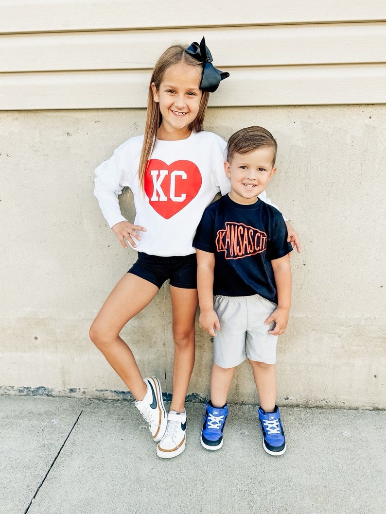 Peut inclure: Deux enfants portant des chemises assorties sur le th&egrave;me de Kansas City. La fille porte un sweat-shirt blanc avec un c&oelig;ur rouge sur lequel est inscrit "KC". Le gar&ccedil;on porte un t-shirt noir avec un contour blanc de l'&eacute;tat du Missouri et les mots "Kansas City" en orange.
