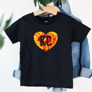 May include: Black t-shirt with a tie-dye heart design and the letters "KC" in the center.