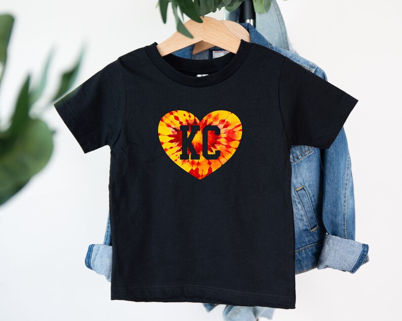 May include: Black t-shirt with a tie-dye heart design and the letters "KC" in the center.