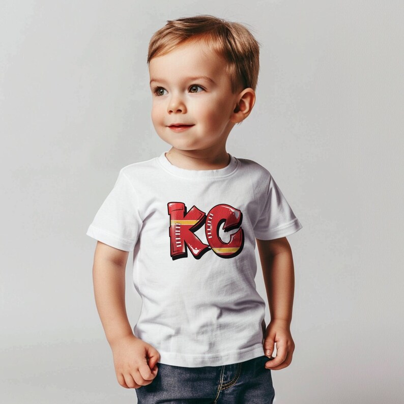 May include: A white t-shirt with a red and yellow graphic of the letters "KC" on the front. A young child is wearing the shirt and looking to the side.