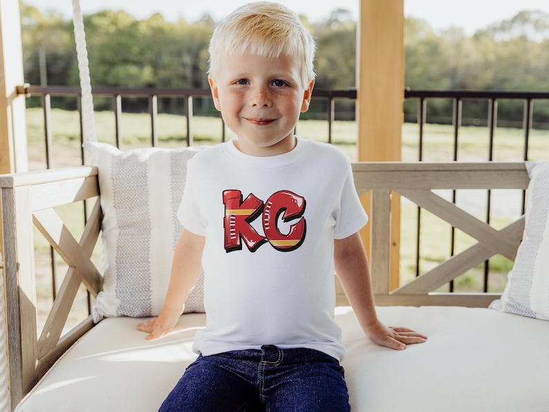 May include: A young child wearing a white t-shirt with a red and yellow "KC" graphic. The child is sitting on a white wooden bench with a gray and white striped pillow.