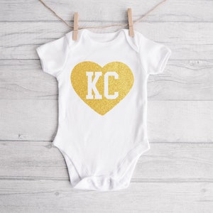 May include: White baby onesie with a gold glitter heart that says "KC" in white letters.
