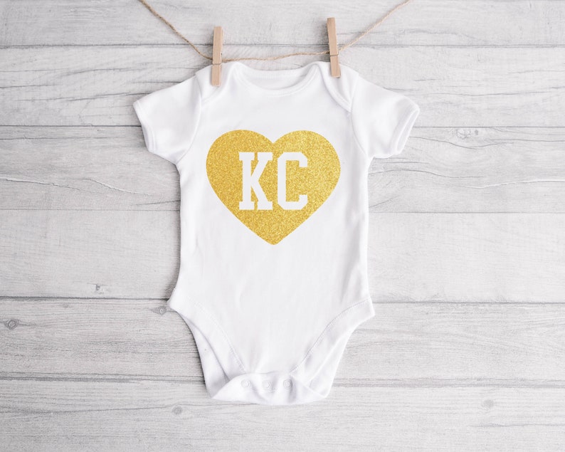 May include: White baby onesie with a gold glitter heart that says "KC" in white letters.