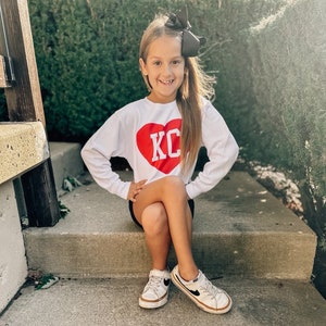 Peut inclure: Une jeune fille portant un t-shirt blanc &agrave; manches longues avec un c&oelig;ur rouge et les lettres "KC" en blanc. Elle porte un n&oelig;ud noir dans les cheveux et des baskets blanches avec des accents noirs.