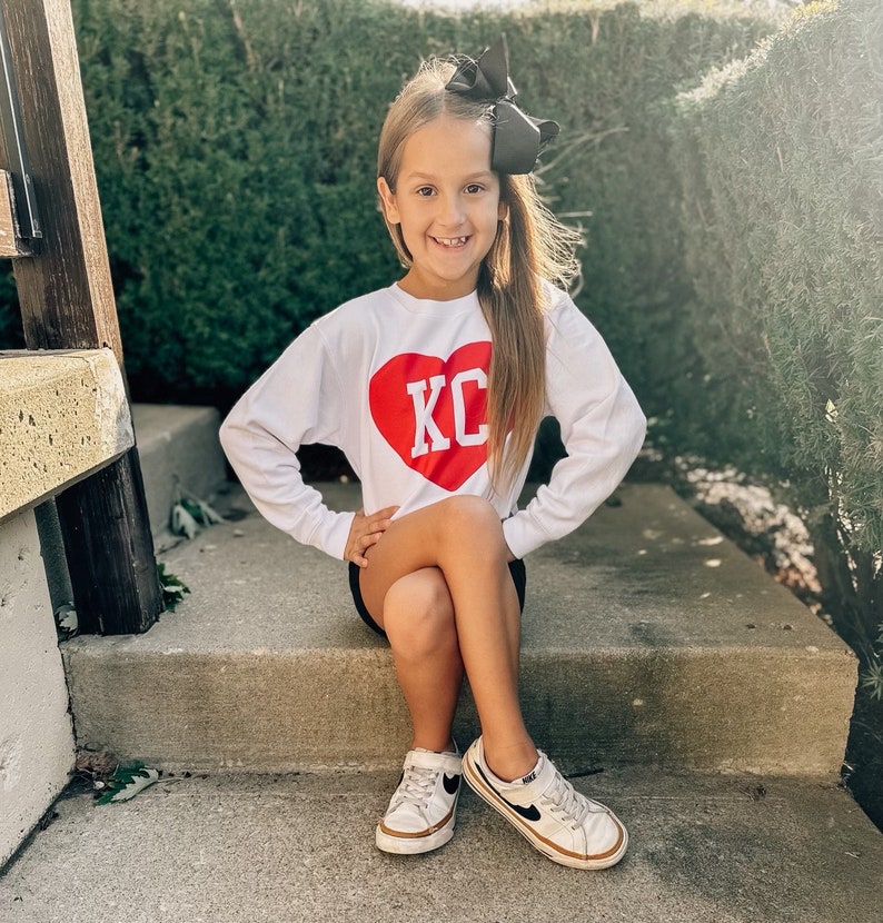Peut inclure: Une jeune fille portant un t-shirt blanc &agrave; manches longues avec un c&oelig;ur rouge et les lettres "KC" en blanc. Elle porte un n&oelig;ud noir dans les cheveux et des baskets blanches avec des accents noirs.