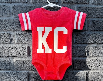 May include: Red baby onesie with white sleeves and white "KC" lettering on the front.