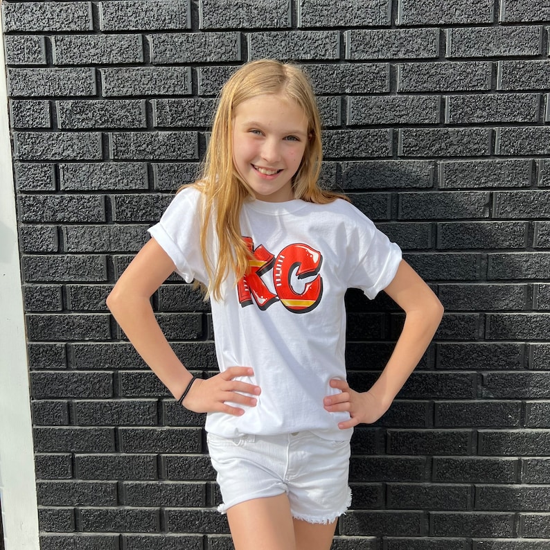 May include: A young person wearing a white t-shirt with a red and gold "KC" graphic. The shirt has short sleeves and a crew neck. The person is standing in front of a black brick wall.