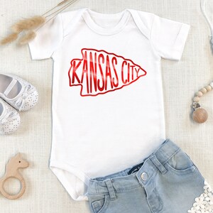 May include: White baby onesie with a red arrow graphic that says "KANSAS CITY". The onesie is paired with light blue denim shorts with white lace trim.