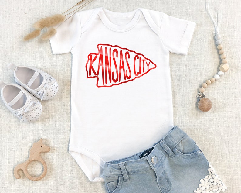 May include: White baby onesie with a red arrow graphic that says "KANSAS CITY". The onesie is paired with light blue denim shorts with white lace trim.