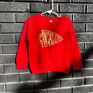May include: Red sweatshirt with a gold arrow design that says "Kansas City" on it. The sweatshirt is hanging on a hook against a brick wall.