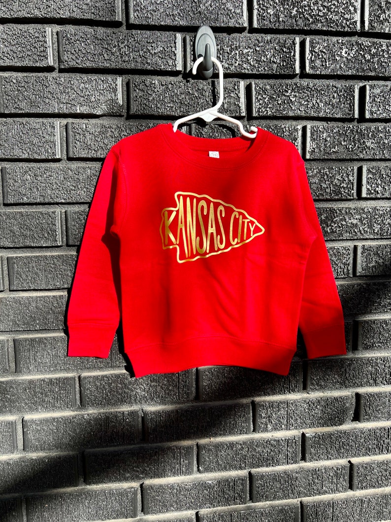 May include: Red sweatshirt with a gold arrow design that says "Kansas City" on it. The sweatshirt is hanging on a hook against a brick wall.