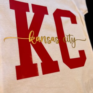 May include: White baby onesie with the letters "KC" in red and the words "kansas city" in gold.