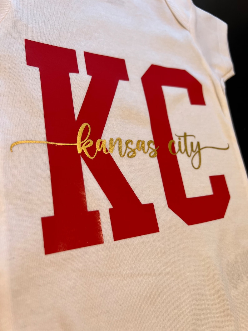 May include: White baby onesie with the letters "KC" in red and the words "kansas city" in gold.