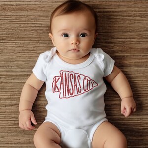 May include: A baby wearing a white onesie with a red "Kansas City" arrow graphic.