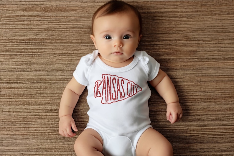 May include: A baby wearing a white onesie with a red "Kansas City" arrow graphic.