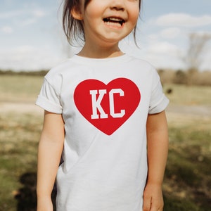 Peut inclure: Un t-shirt blanc avec un c&oelig;ur rouge sur lequel sont inscrites les lettres "KC" en blanc.
