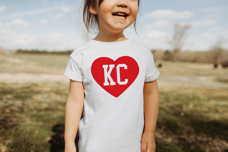 Peut inclure: Un t-shirt blanc avec un c&oelig;ur rouge sur lequel sont inscrites les lettres "KC" en blanc.