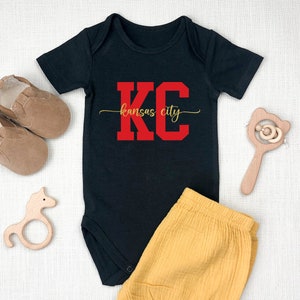 May include: Black baby onesie with red "KC" lettering and gold "Kansas City" text. The onesie is paired with yellow shorts.