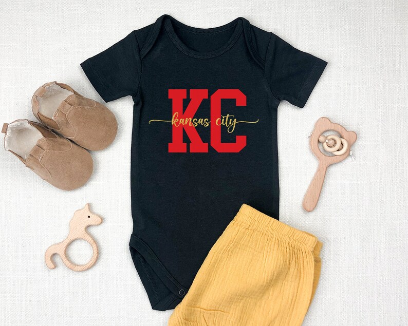 May include: Black baby onesie with red "KC" lettering and gold "Kansas City" text. The onesie is paired with yellow shorts.