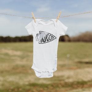 May include: White baby onesie with a black and white arrow design that says "KANSAS CITY" on it. The onesie is hanging on a clothesline.