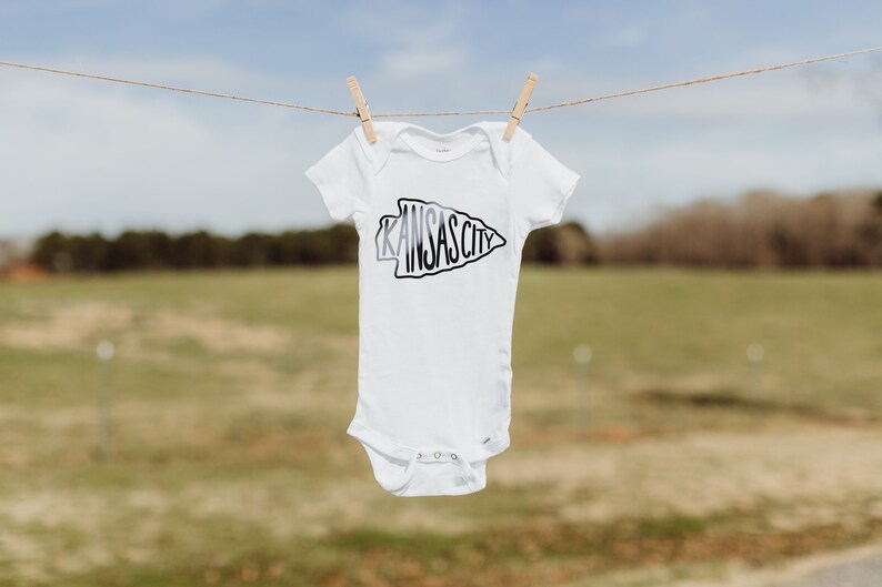 May include: White baby onesie with a black and white arrow design that says "KANSAS CITY" on it. The onesie is hanging on a clothesline.