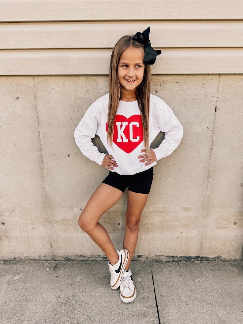 Peut inclure: Une jeune personne portant un sweat-shirt blanc avec un c&oelig;ur rouge et les lettres "KC" en blanc. Elle porte &eacute;galement un short noir et des baskets blanches avec des accents noirs.