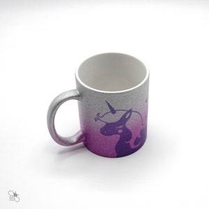 Könnte beinhalten: Eine weiße Keramiktasse mit einem rosa und silbernen Glitzer-Verlauf. Die Tasse zeigt eine violette Silhouette eines Einhorns mit einem Horn.