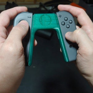 May include: A green 3D printed grip attachment for a Nintendo Switch controller. The grip features the Super Smash Bros. logo and is designed to make the controller more comfortable to hold.